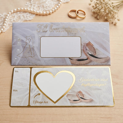 Et bryllupskort med et hvitt felt i hjerte. Skriv inn gaven du vil gi og fest over skrapelappen som følger med produktet. Perfekt til alle slags gaver, uansett om du gir penger, en opplevelsegave, eller noe helt annet. Gullfoliert tekst og kanter. Bak kortet er en elegant konvolutt som følger med produktet. Kort og konvolutt er håndlaget av Hjertelig AS.