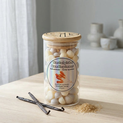Duftvoks i glass på et bord. Ved siden av vanlijestang og brunt sukker, som illustrerer duften til nostalgiske kandispinner. Håndlaget i norge med soyavoks og bivoks. Hvert glass inneholder 90g voks.