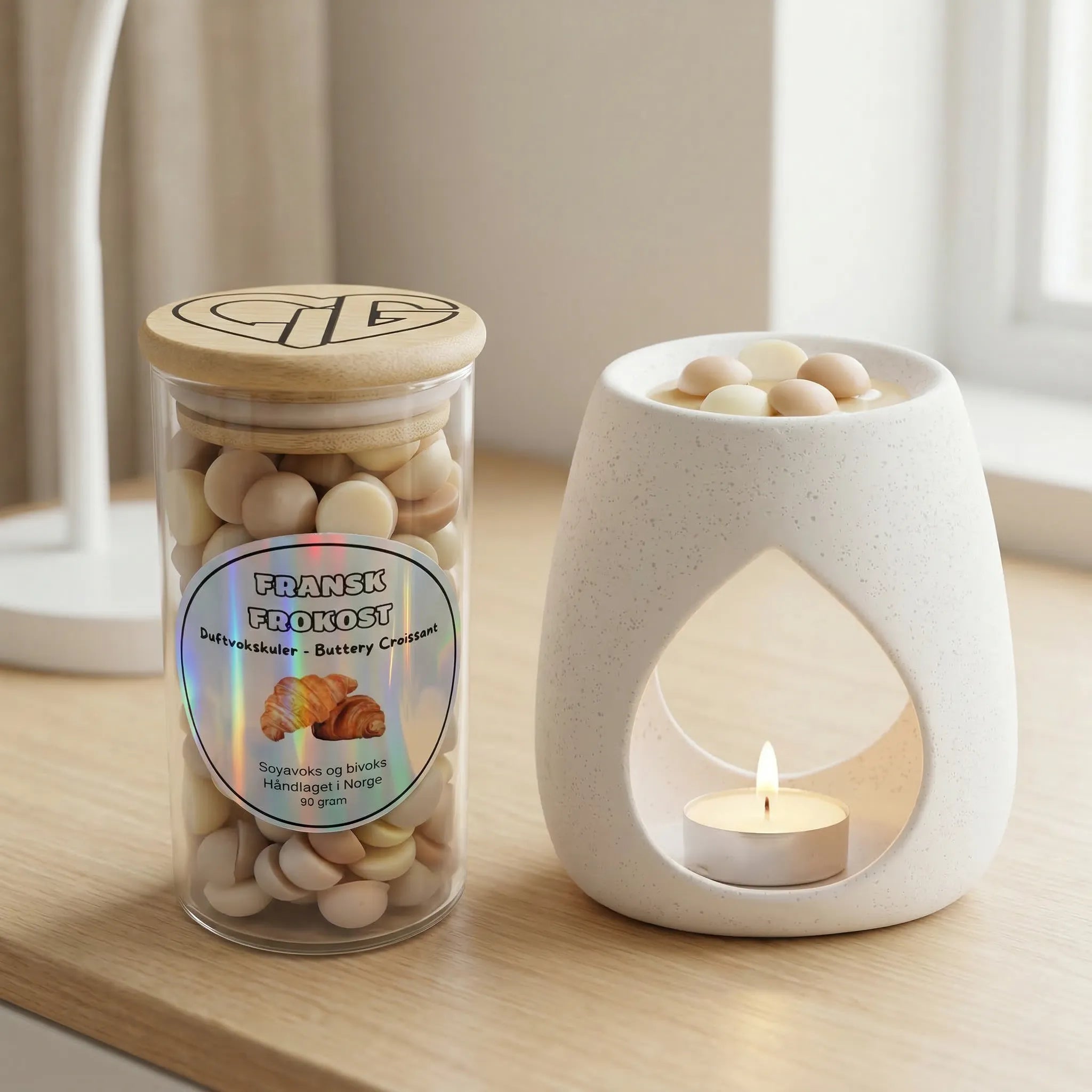 Duftvokskuler i glass ved siden av en duftbrenner med telys. Glasset inneholder 90g duftvoks som er laget av soyavoks og bivoks. Fransk frokost fordi dufrvoksen lukter som nystekte croissanter. Duftbrenneren er fylt med noen vokskuler, som smelter og sprer herlig duft i rommet. Håndlaget i norge.