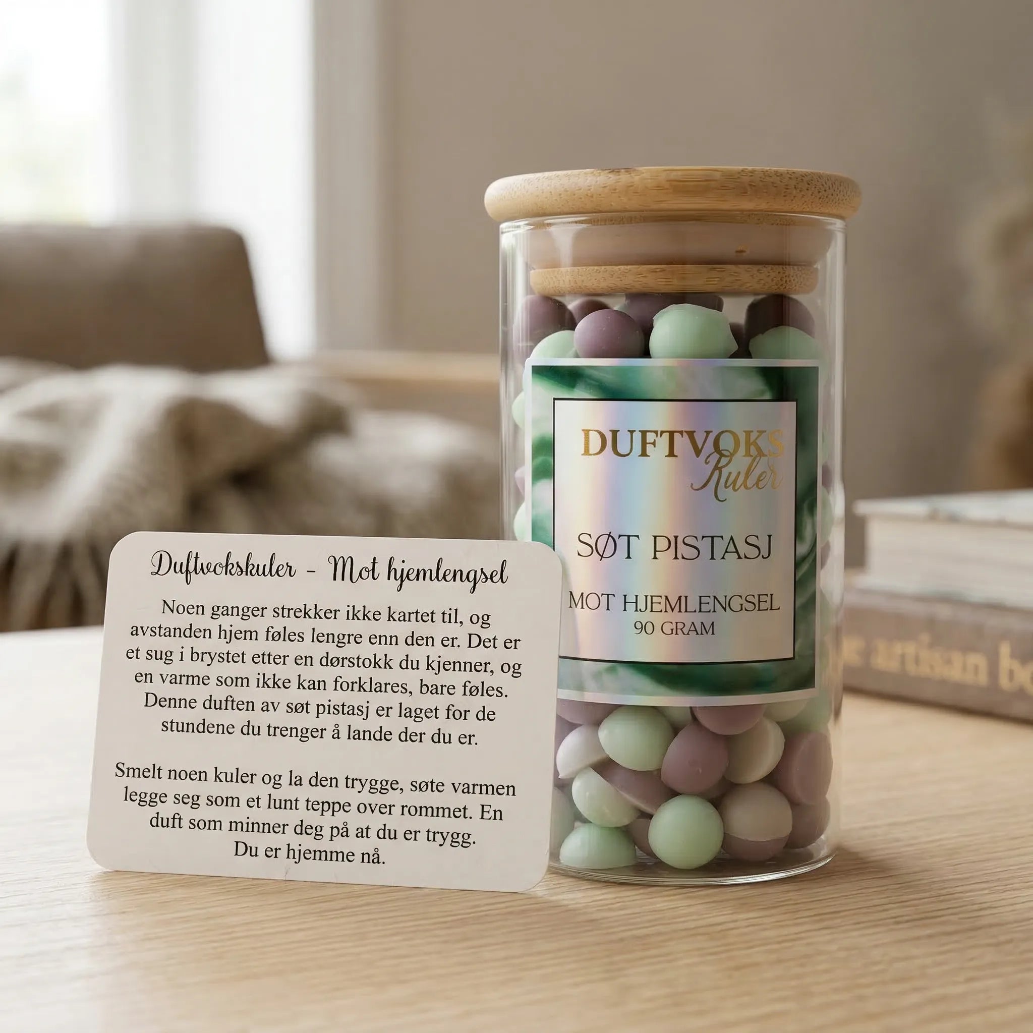 Duftvoks mot hjemlengsel. Duftvokskuler som lukter søt pistasj. Et glass med 90g voks og et følelseskort som følger med produktet står på et bord. Håndlaget i Norge med bivoks og soyavoks. Gullfoliert etikett