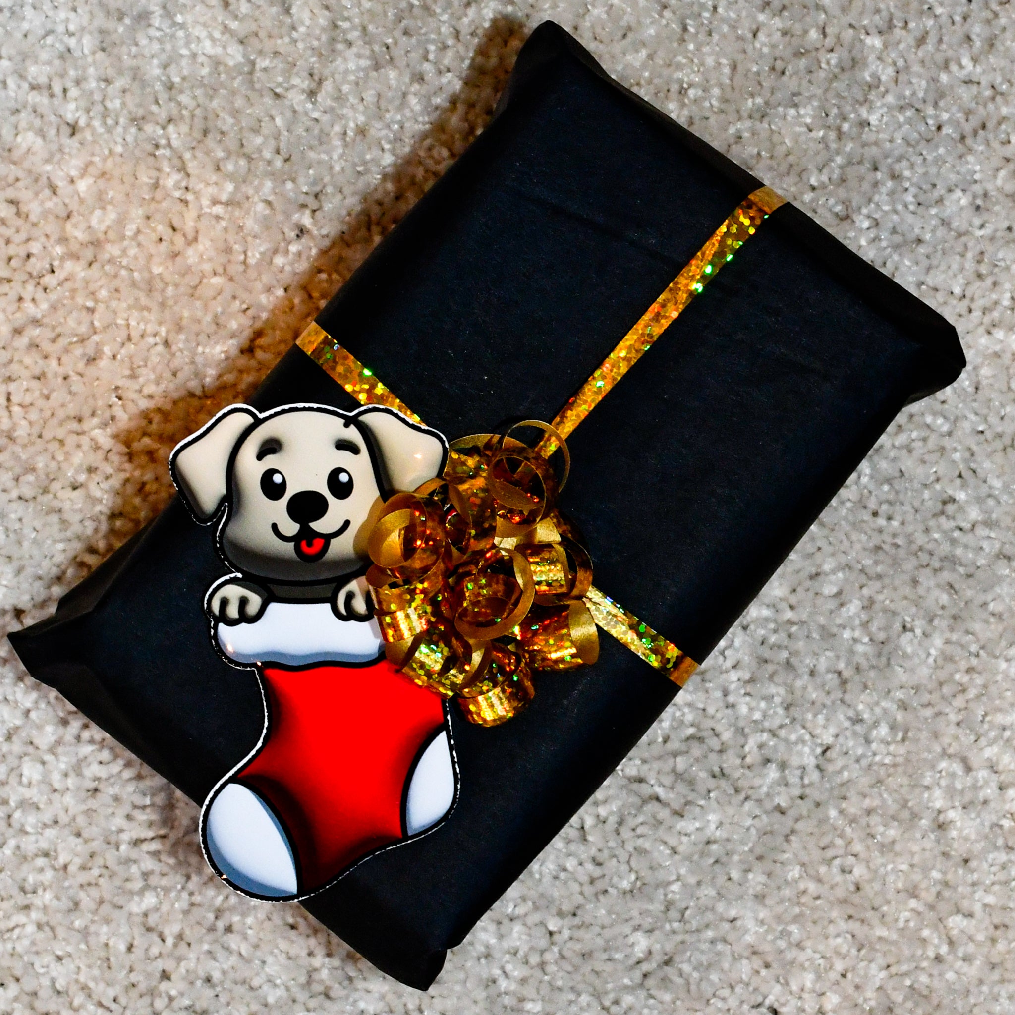 Julegave som er pakket inn med gull gavebånd og har en merkelapp til og fra navnelapp på seg. Navnelappen er festet i gavebåndet og er en søt liten valp som sitter i en julestrømpe. Det ser ut som at den stikker litt ut av papiret da den er i et boble design. Nydelig detalje til julegavene. Gøy for store og små og gir skikkelig julestemning til gavene. 