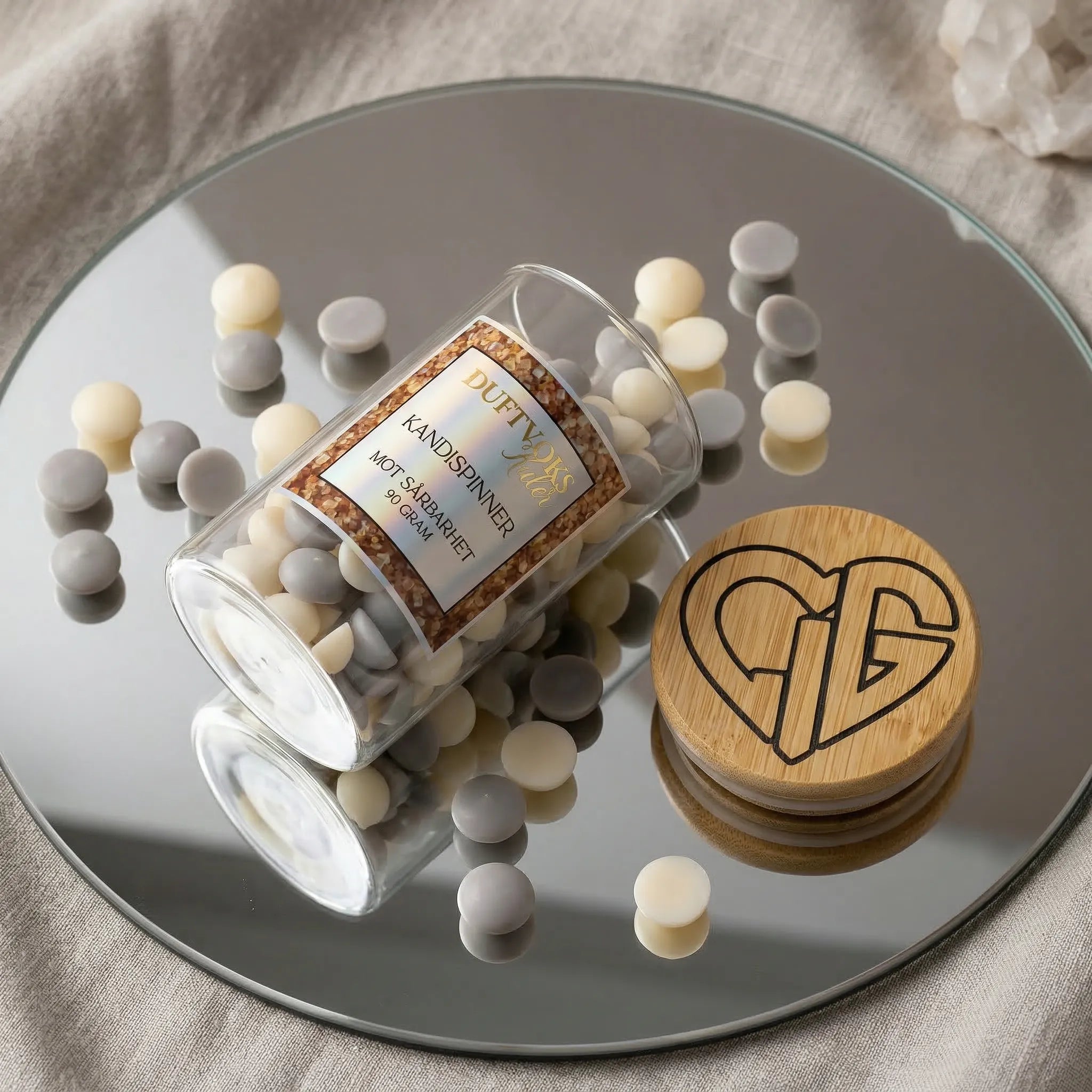 Små duftvokskuler i glass som ligger på et speil. Trelokk med Hjertelig AS sin logo ved siden av. Duftvoksen er laget av bivoks og soyaboks, og lukter kandispinner. Denne søte duften skal virke lindrende mot sårbarhet. Håndlaget i Norge.