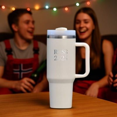 Russ 2025 termokopp med lasergravering som står på et bord. Termokoppen er i beige farge. I bakgrunnen sitter det to russ som smiler og har det gøy. Årets russemerch 2025 fra Hjertelig.no