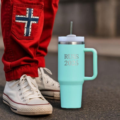 En turkis termokopp med RUSS 2025 lasergraver. Ved siden av står det en russ med hvite sko og rød russebukse. Termokoppen er årets russemerch hos Hjertelig.no