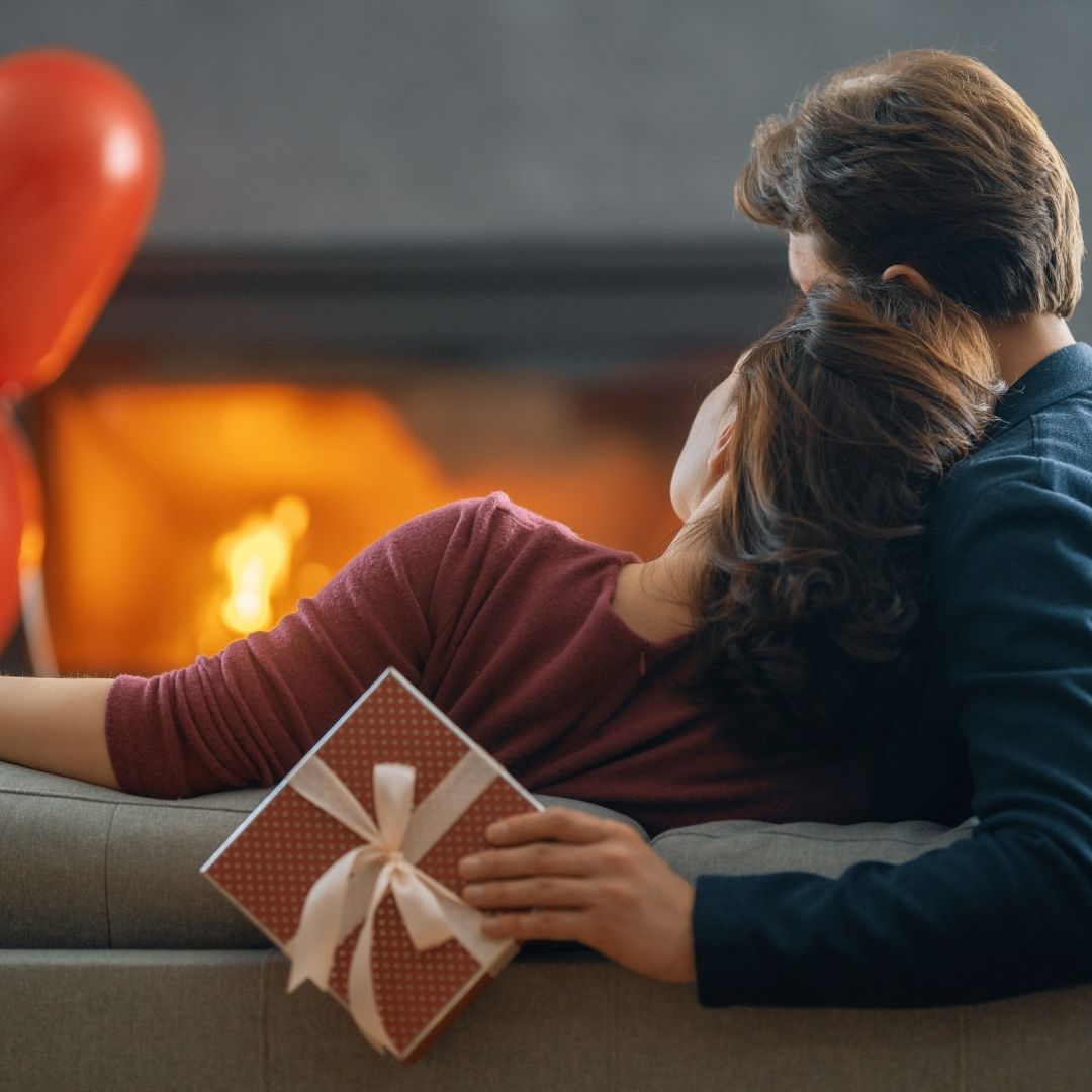 Et kjærestepar som sitter i sofaen, der mannen har gjemt en gave bak ryggen på kvinnen. Bildet illustrerer Valentine