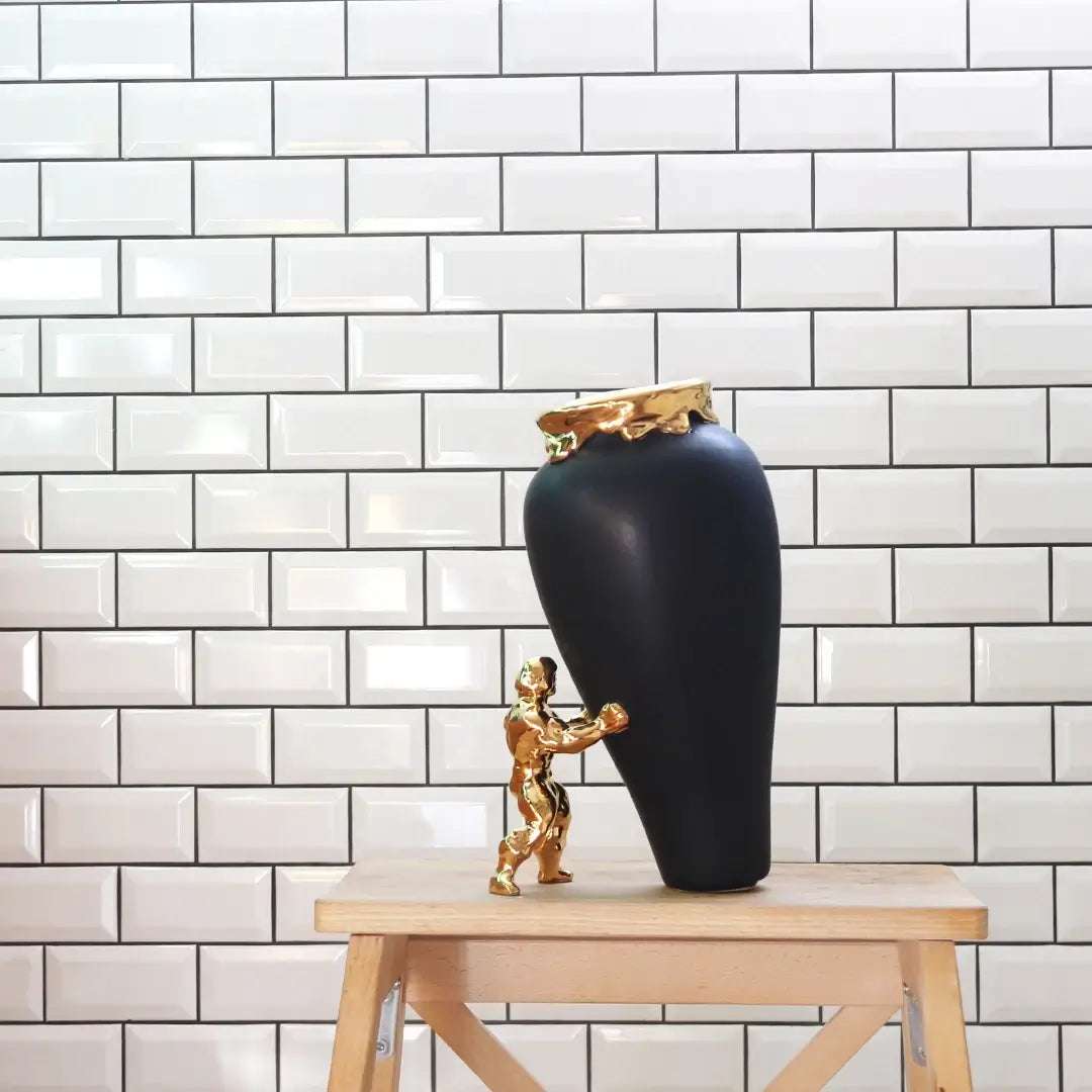 En sort vase med gullkant på et lite bord. En superhelt i gull støtter opp vasen slik at den ikke faller. Ekte kunst og blomstervase som håndlages i nederland. Et blikkfang av en vase som virkelig gir rommet et løft.